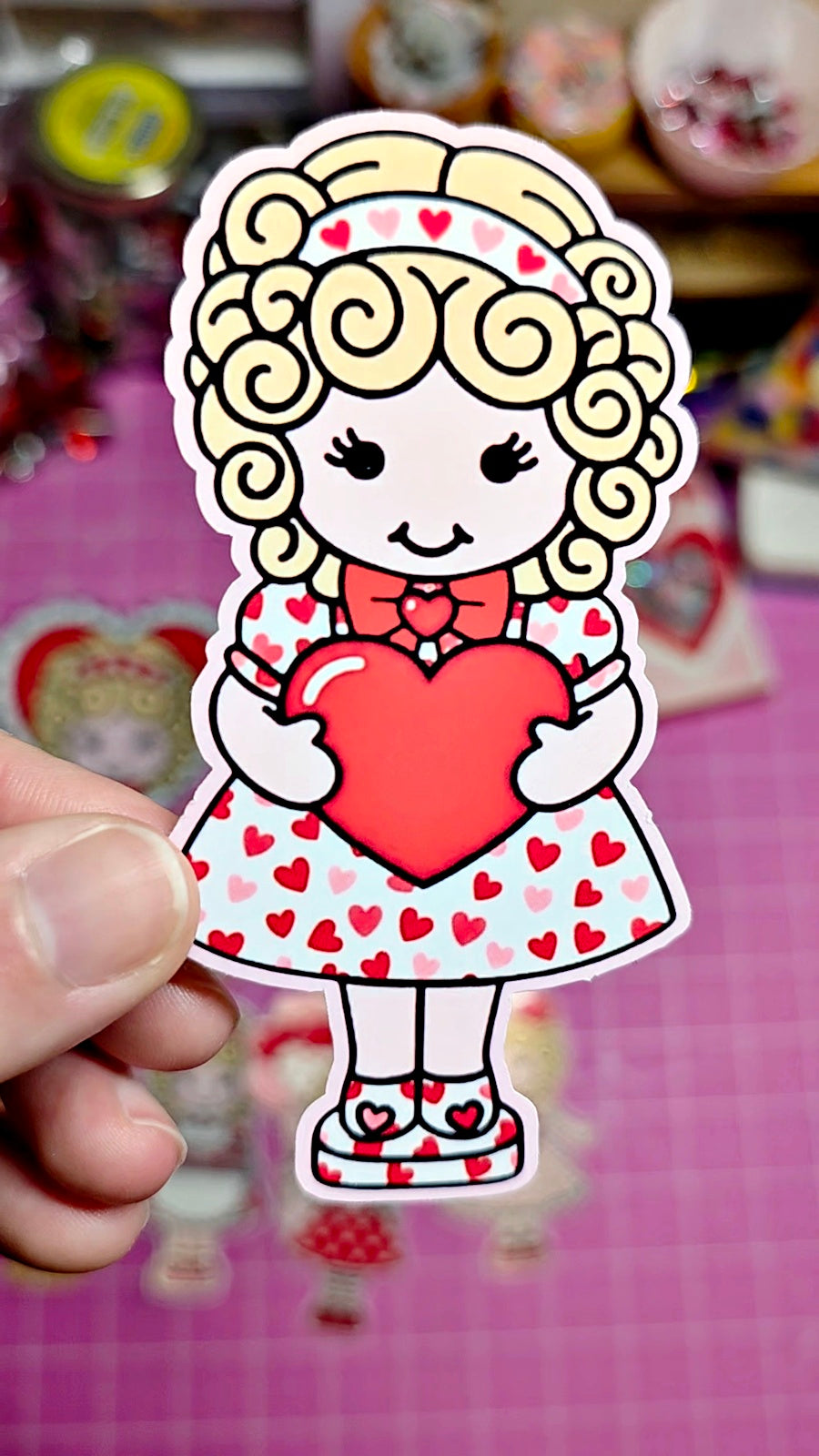 Sticker of a cartoon character holding a heart, held by a hand on a colorful background.