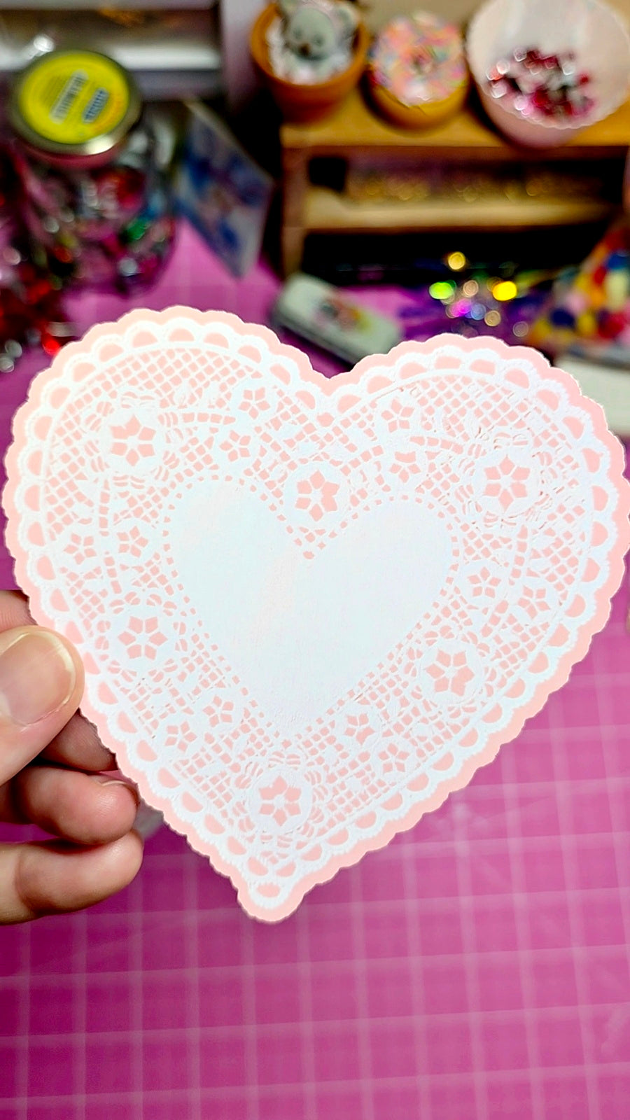 Heart-shaped pink doily held by a hand on a pink surface with blurred background