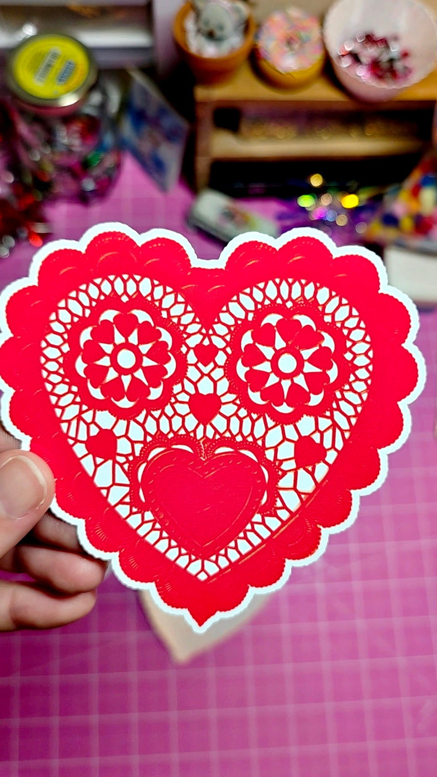 Red heart-shaped cutout with intricate patterns held by a hand on a pink checkered surface.