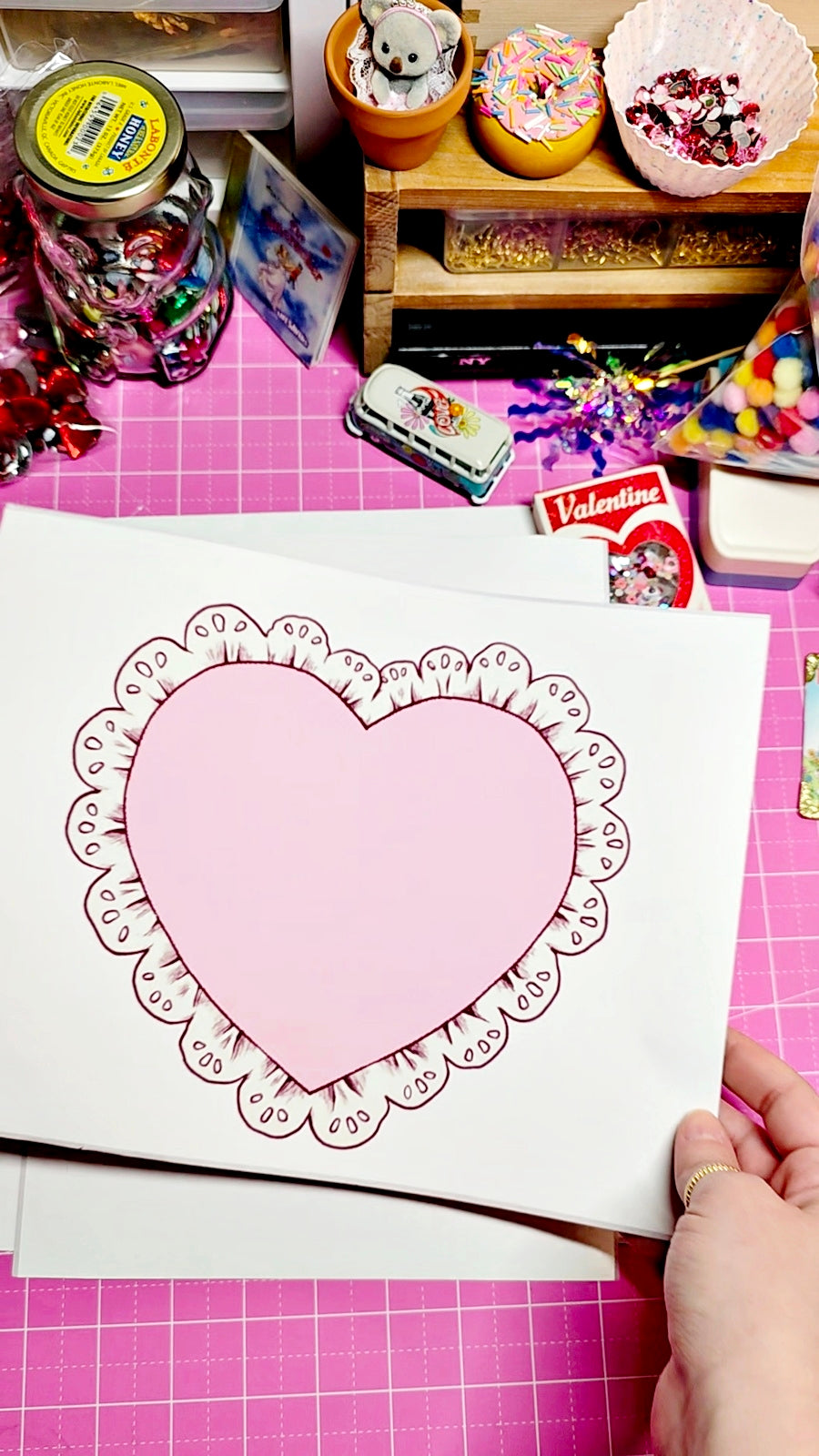 Heart-shaped paper cutout on a pink cutting mat with craft supplies in the background