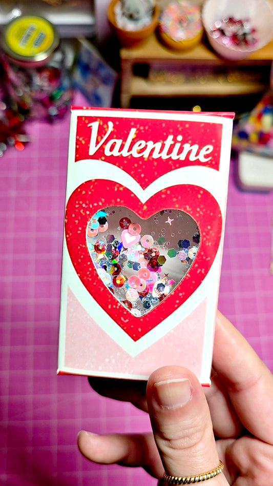 Valentine's Day card with heart cutout filled with colorful sequins, held by a hand on a pink background.