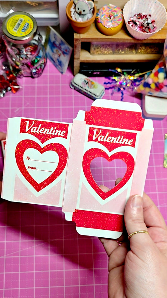 Valentine's Day card with heart design held by a hand on a pink surface with toys in the background.