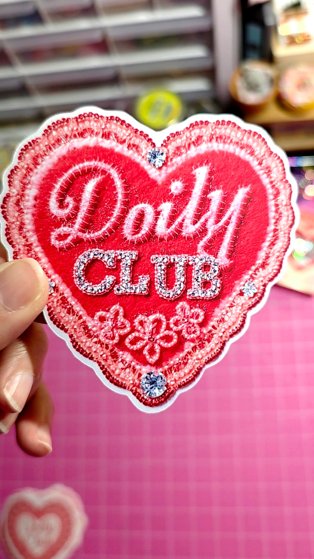 Heart-shaped patch with 'Doily Club' text held by a hand on a pink background