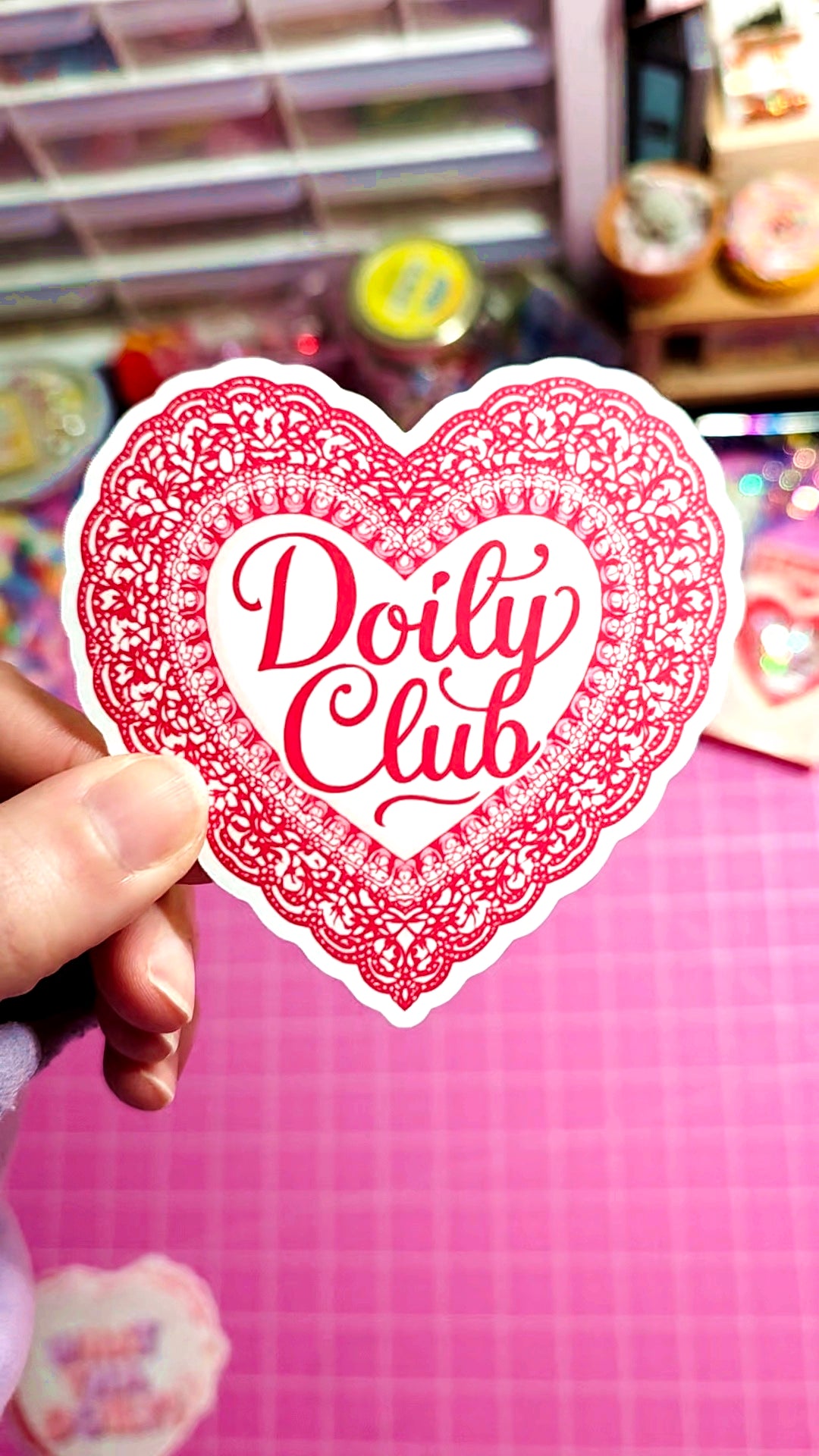 Heart-shaped sticker with 'Doily Club' text held by a hand against a colorful background.