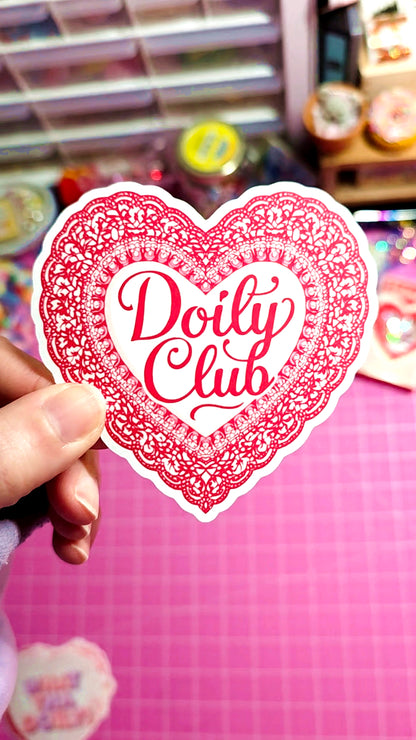 Heart-shaped sticker with 'Doily Club' text held by a hand against a colorful background.