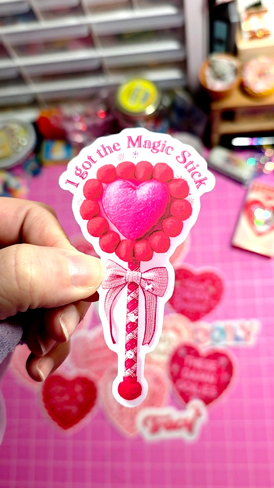 Sticker of a pink heart with candy cane stick and 'I got the magic stick' text, held by a hand against a colorful background.