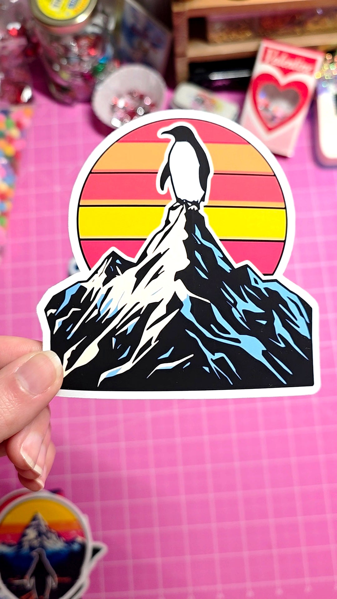 Sticker of a penguin on a mountain with a sunset background held over a pink grid background.