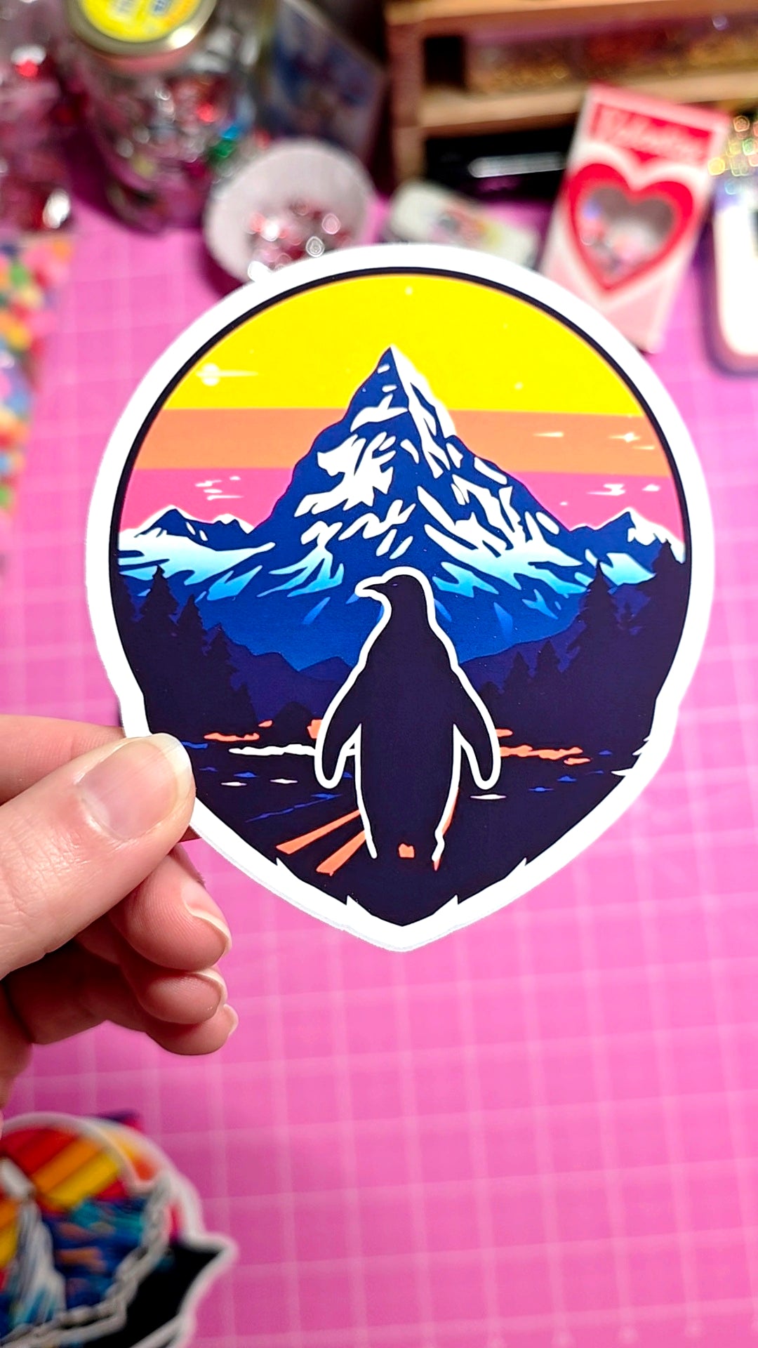 Sticker of a mountain and penguin silhouette with a colorful sky held against a pink background.