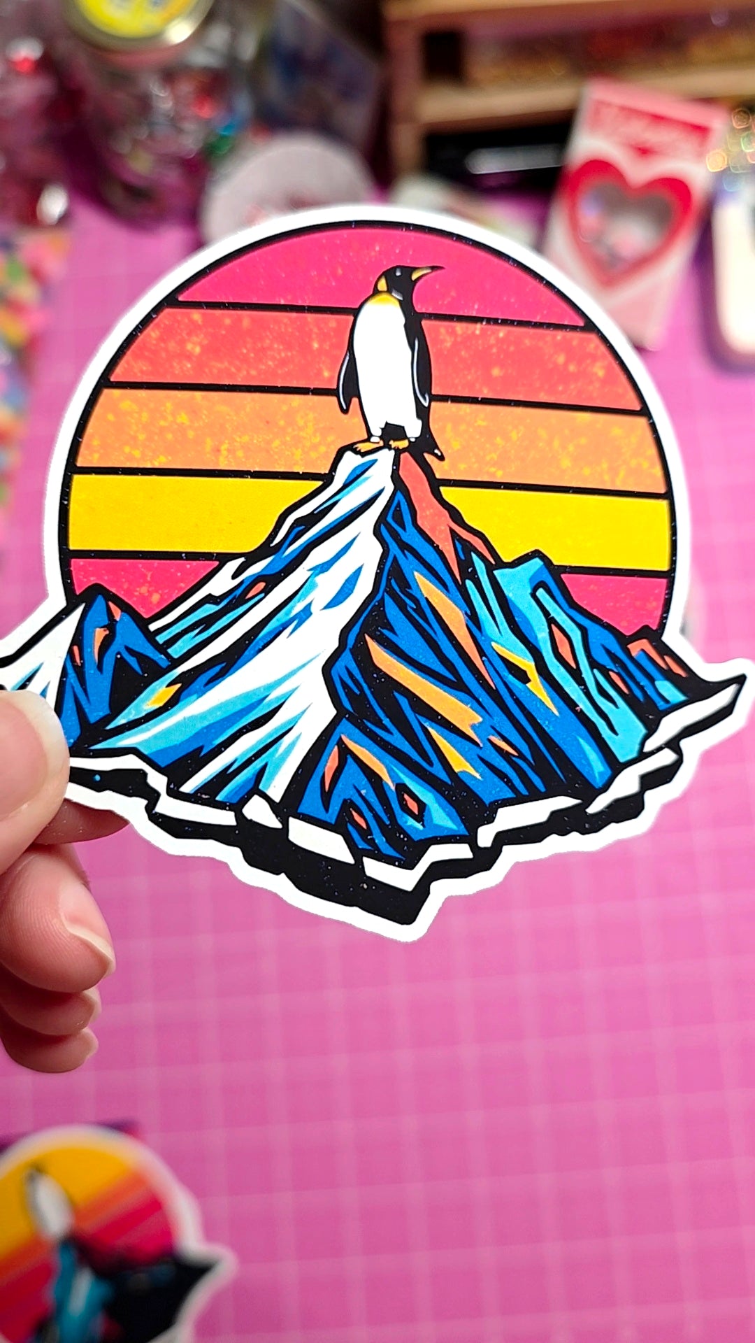 Sticker of a penguin on a mountain with a sunset background held against a pink grid background.