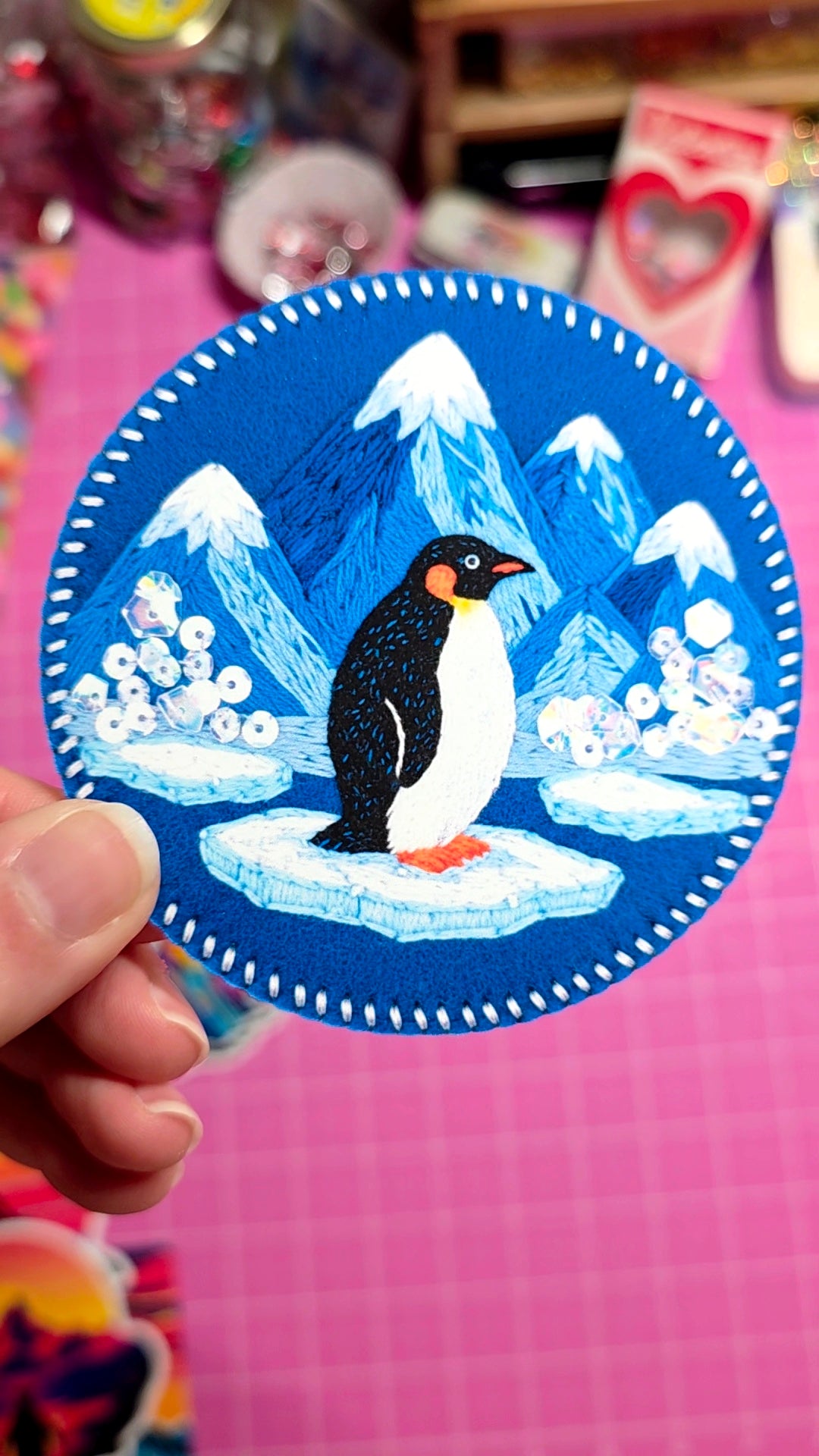 Sticker of a penguin on an ice floe with mountains in the background, held against a pink grid background.