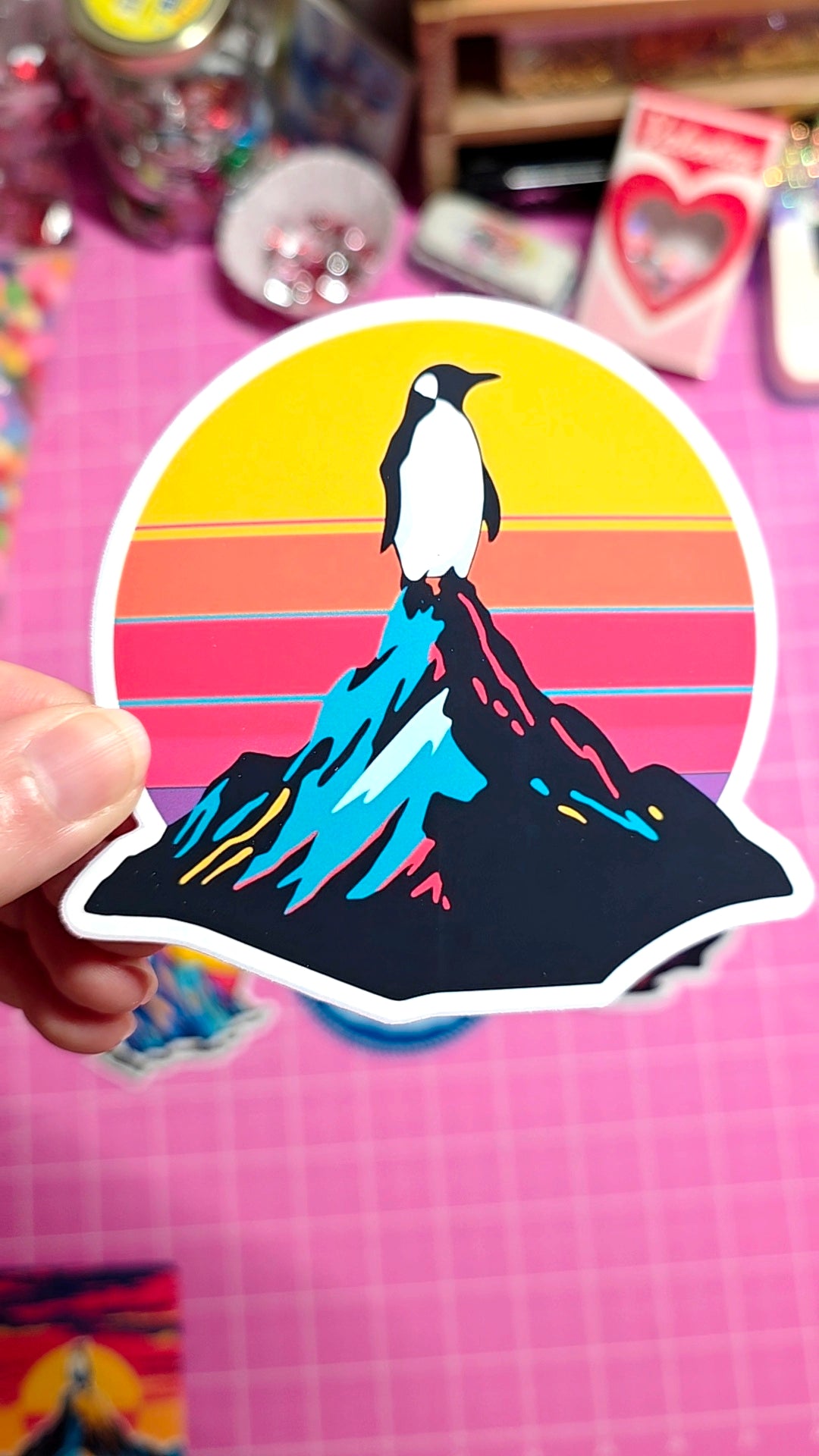 Sticker of a penguin on a mountain with a colorful sunset background held by a hand on a pink grid surface.