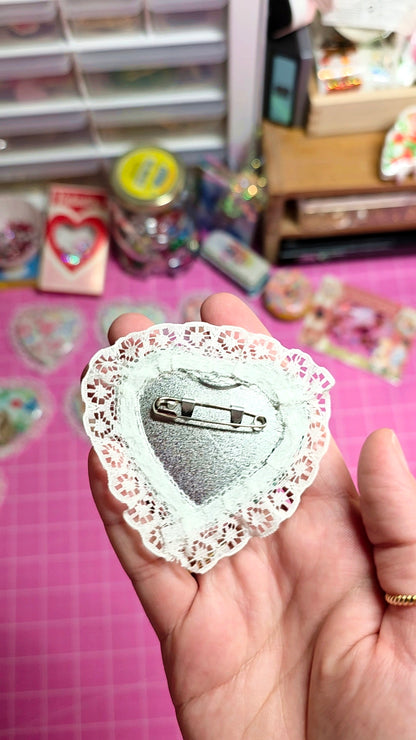 Heart-shaped brooch with lace detailing held in a hand on a pink surface with craft supplies.
