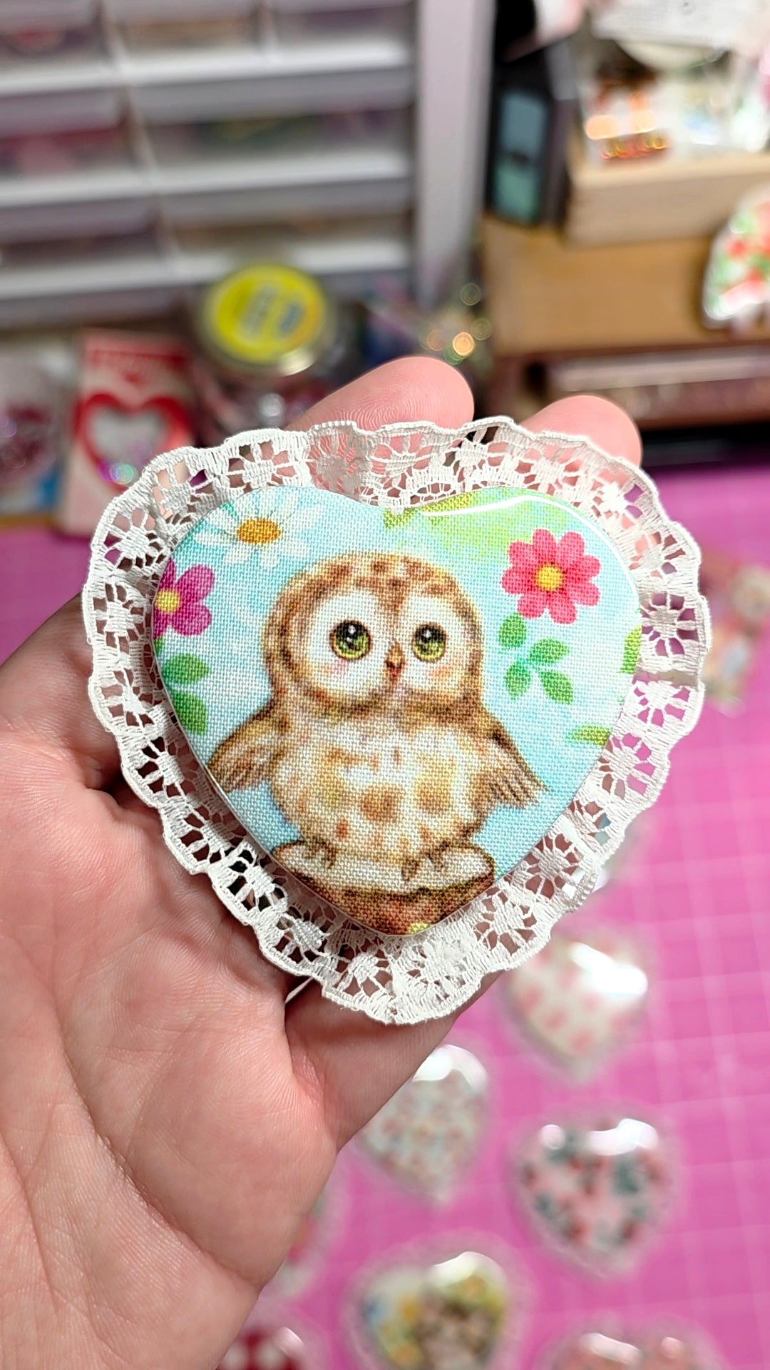 Heart-shaped decorative sticker with an owl design held by a hand on a pink surface.