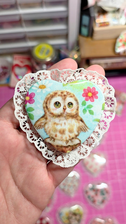 Heart-shaped decorative sticker with an owl design held by a hand on a pink surface.