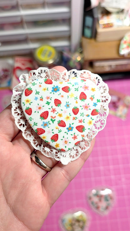 Heart-shaped paper cutout with strawberry pattern held by a hand against a blurred background