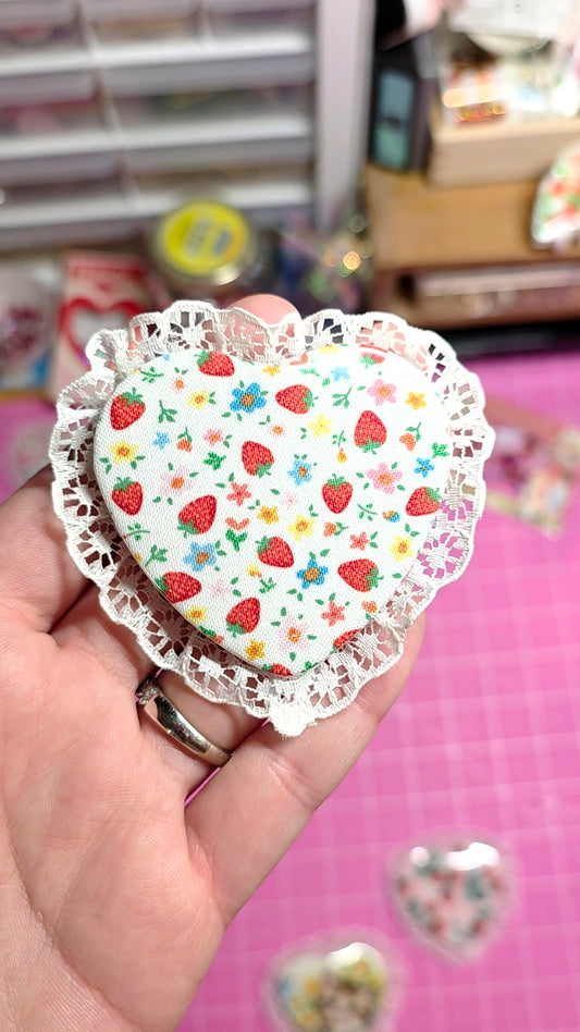 Heart-shaped paper cutout with strawberry pattern held by a hand against a blurred background