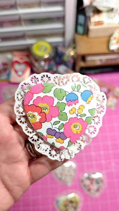 Heart-shaped paper cutout with colorful floral design held by a hand on a pink surface.