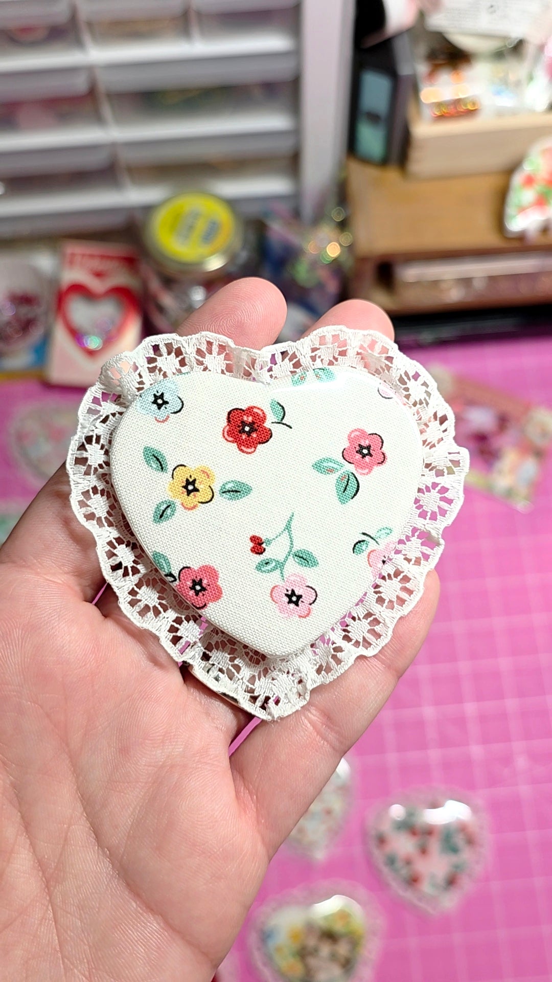 Heart-shaped decorative item with floral patterns held in a hand against a pink background.