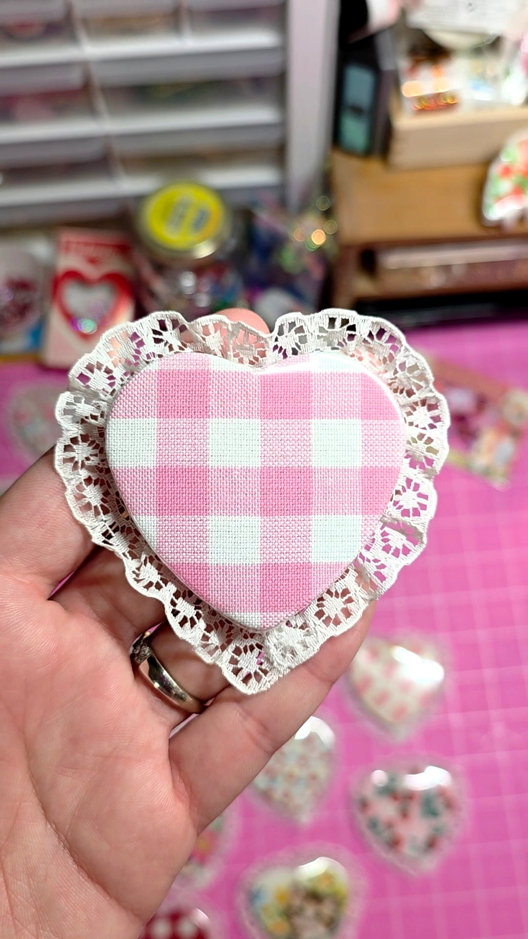 Heart-shaped pink and white checkered fabric with lace trim held by a hand on a pink background.