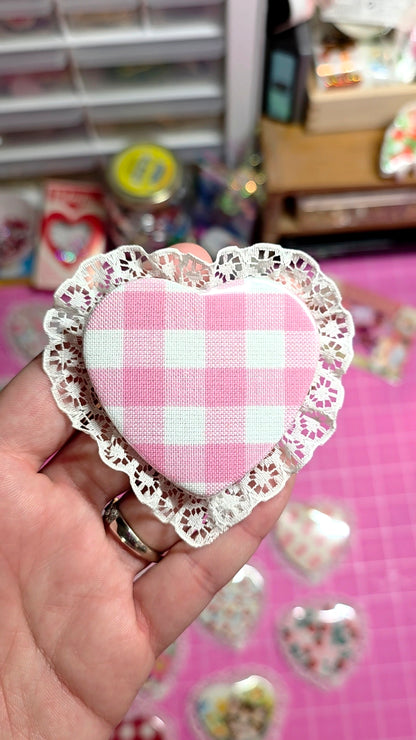 Heart-shaped pink and white checkered fabric with lace trim held by a hand on a pink background.