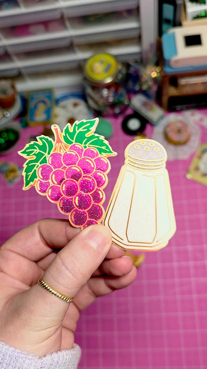 Hand holding a decorative item with grapes and a saltshaker design against a colorful background.