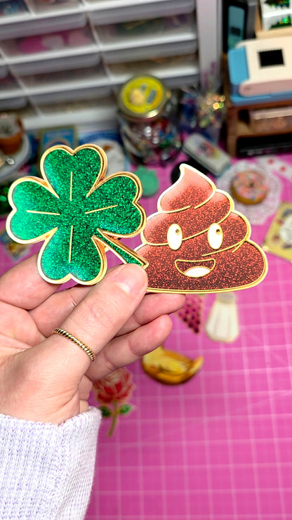 Hand holding a green glittery four-leaf clover and a pink poop emoji sticker against a colorful background.