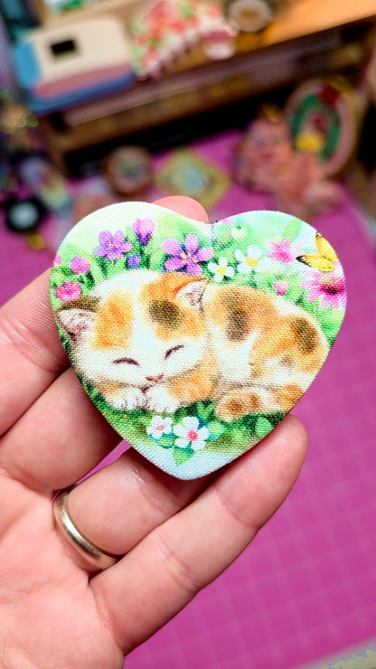 Heart-shaped patch with a cat design held by a hand on a pink surface.