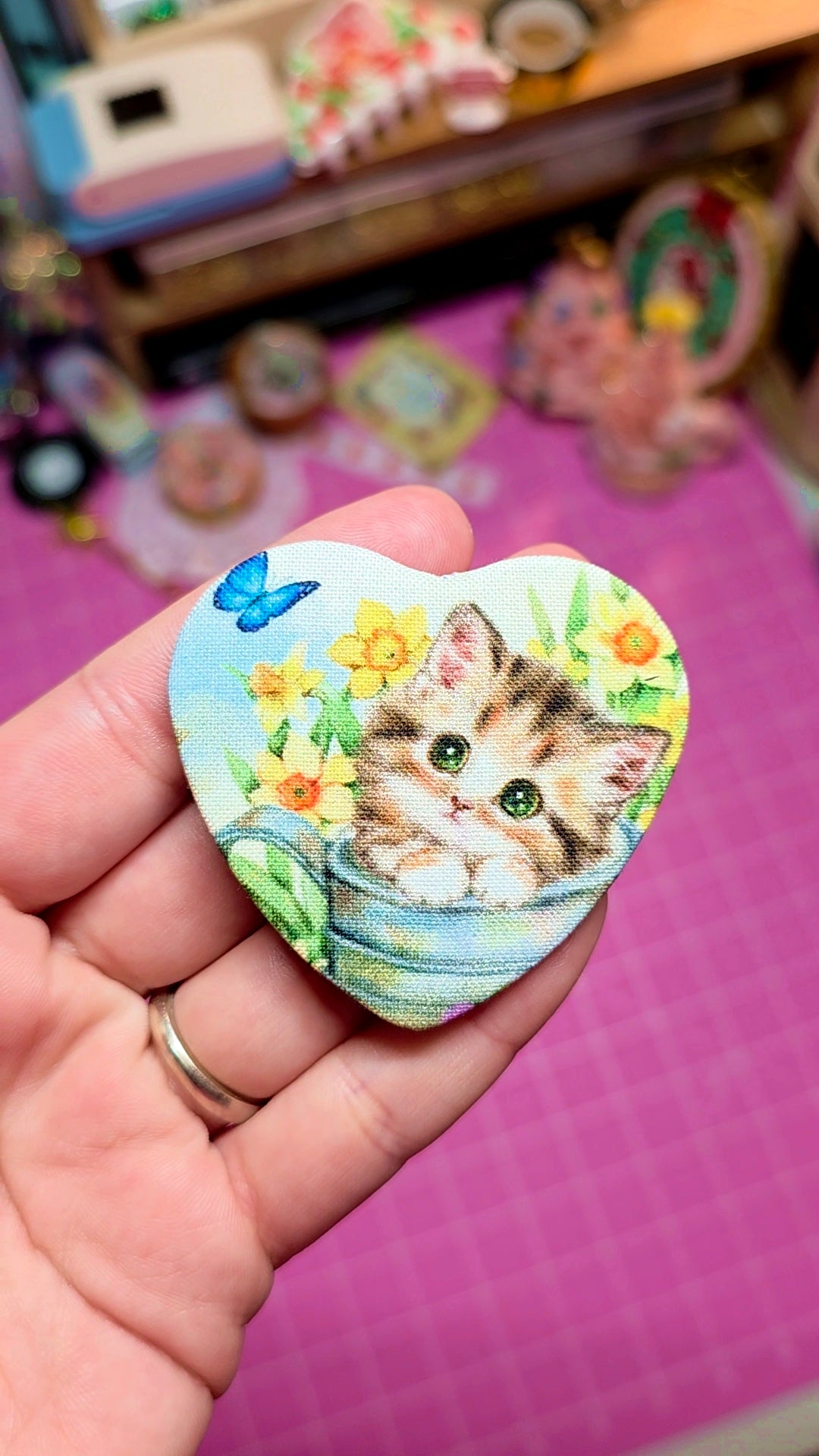 Heart-shaped magnet with a cat illustration held by a hand on a pink surface.