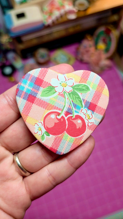 Heart-shaped decorative item with cherry design held in a hand against a colorful background