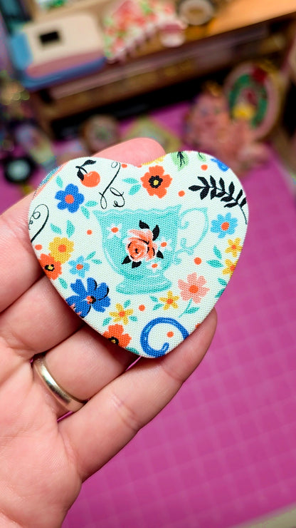 Heart-shaped decorative item with floral patterns and a teapot design held by a hand on a pink surface.