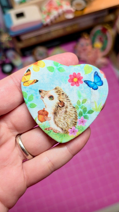 Heart-shaped pin with a hedgehog design held in a hand against a colorful background.
