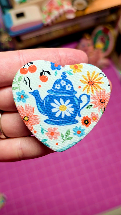Heart-shaped decorative sticker with a blue teapot design held by a hand on a pink surface.