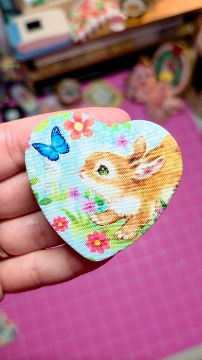 Heart-shaped patch with a bunny and butterfly design held in a hand against a pink background.
