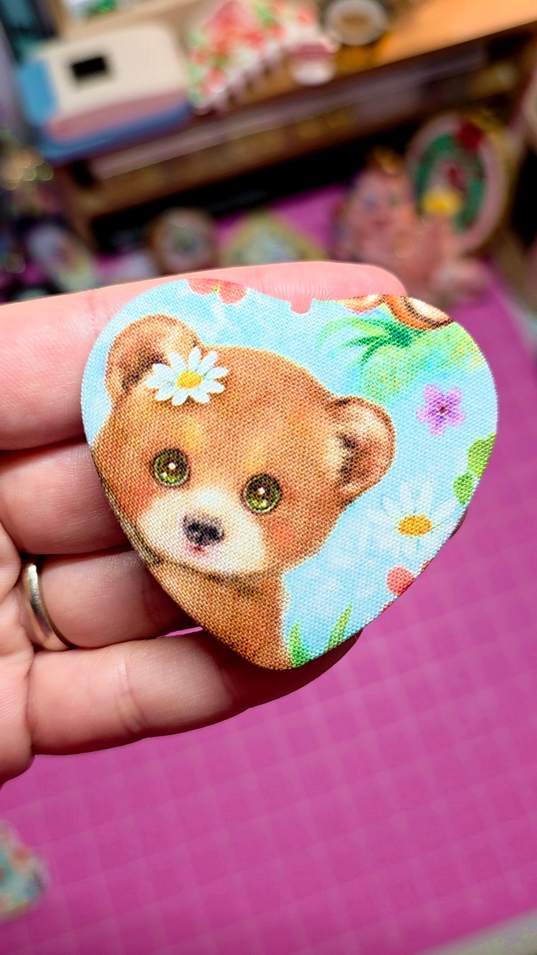 Fabric patch of a bear with flowers held by a hand on a pink surface.