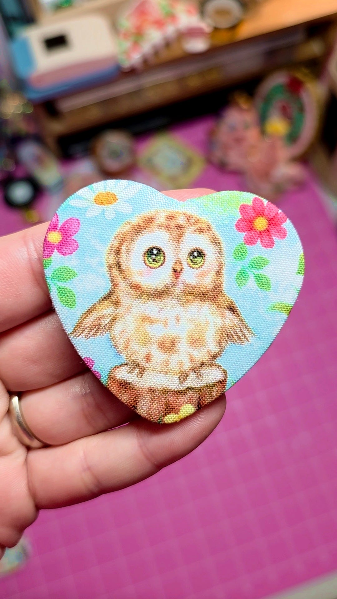 Heart-shaped sticker with an owl design held by a hand on a pink surface.