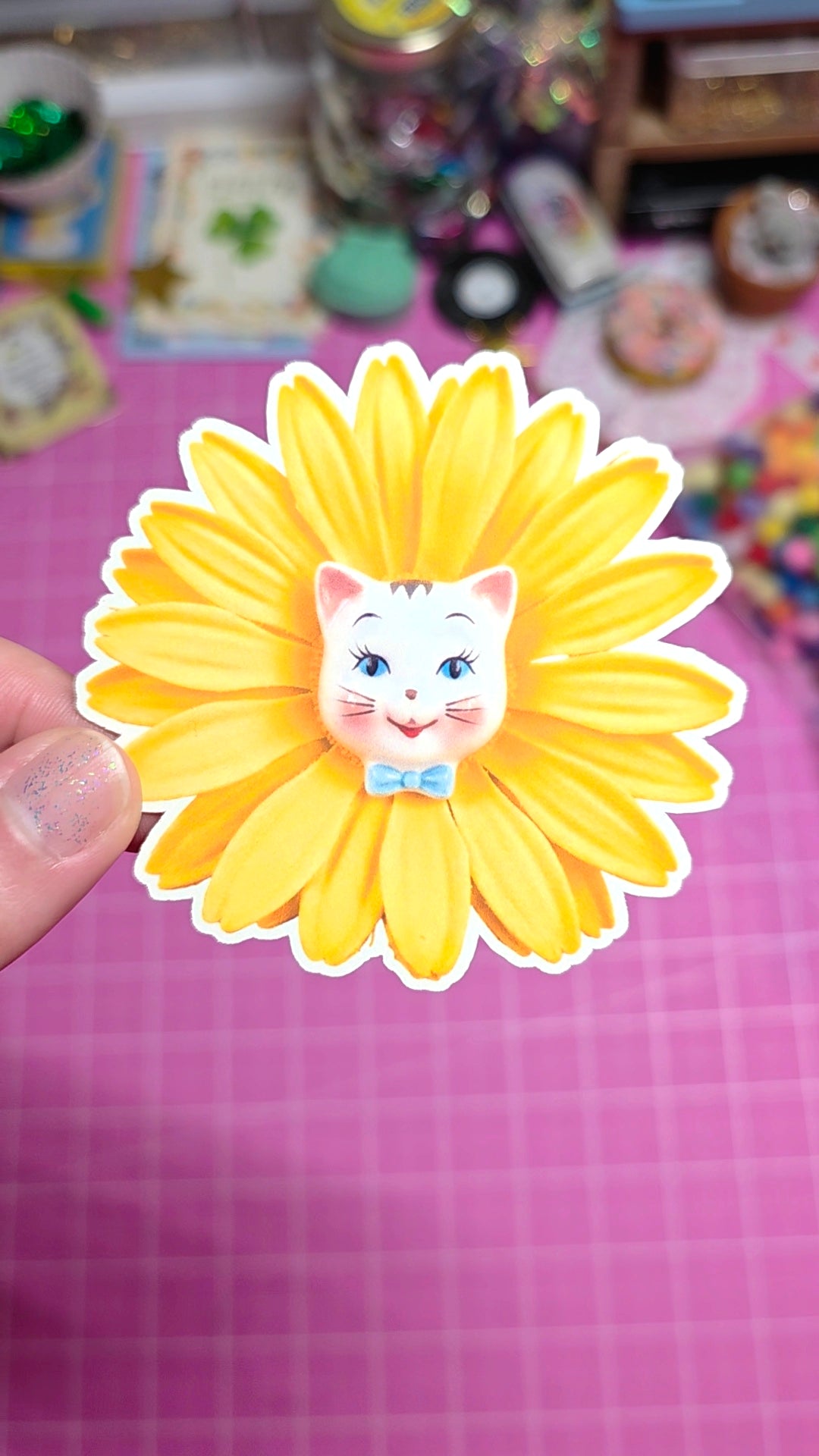 Yellow flower-shaped sticker with a cat face design held by a hand on a pink checkered background.