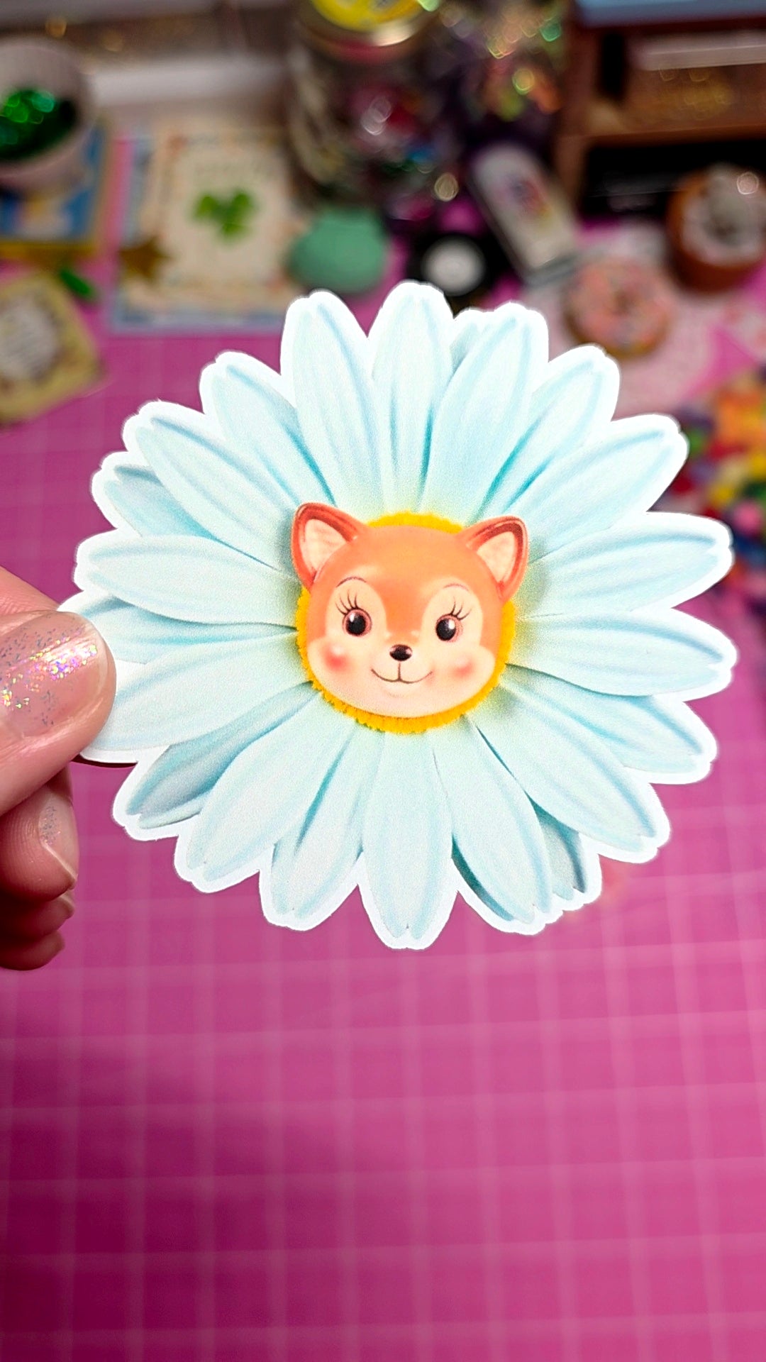 Flower-shaped sticker with a fox design held against a pink grid background