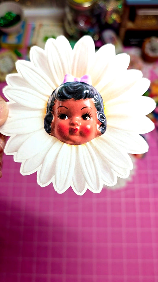 Vintage-style toy head with black hair and red lips, surrounded by a white flower on a pink checkered background.