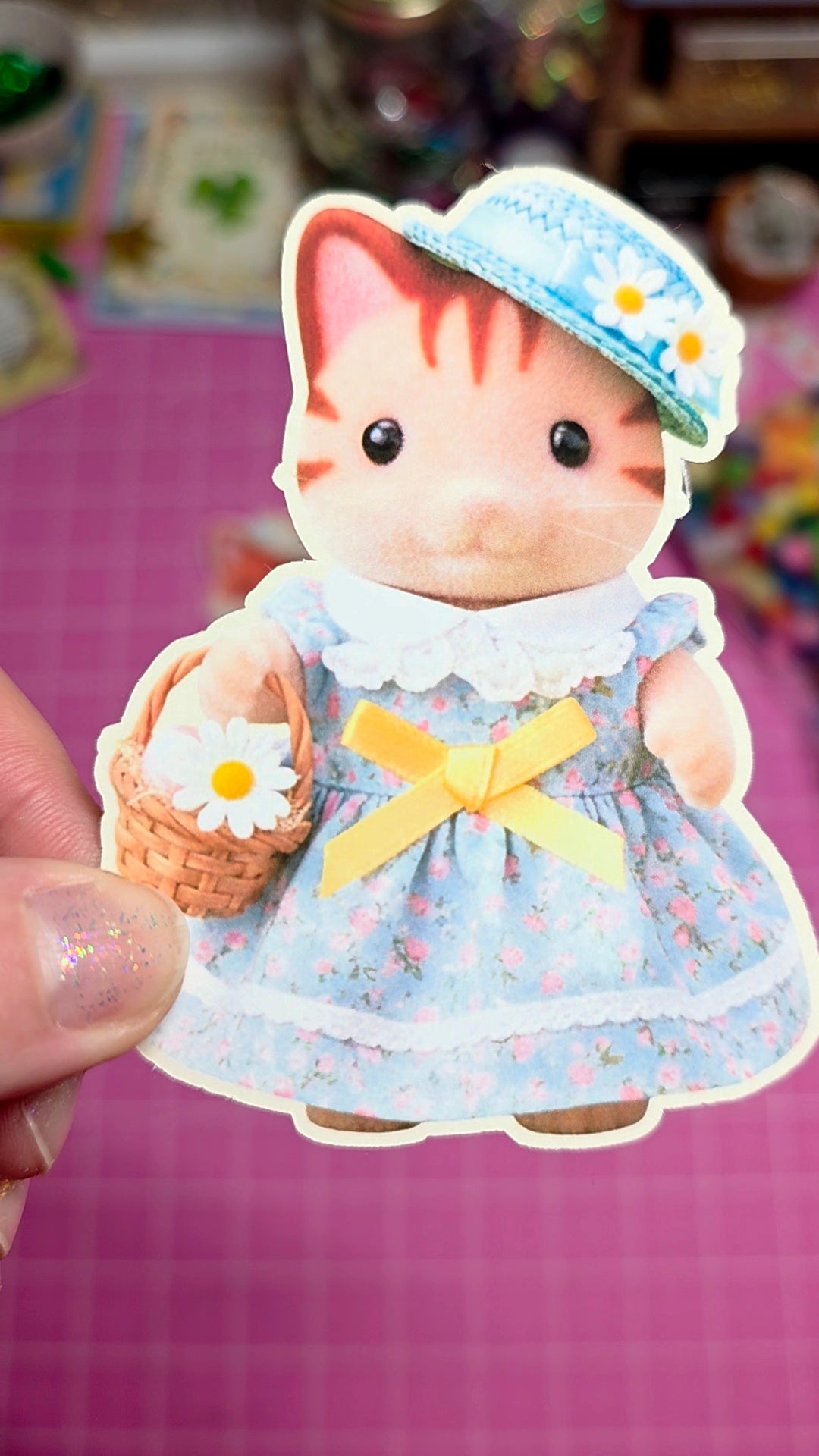 Small figurine of a cat in a blue dress and hat holding a basket with a flower, on a pink surface.