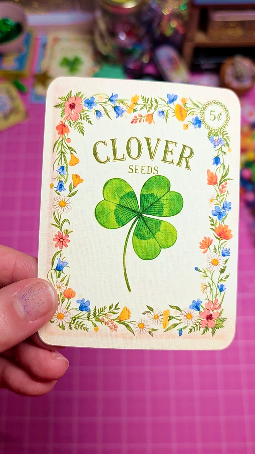 Card with a green clover and floral design, labeled 'Clover Seeds', held by a hand on a pink background.