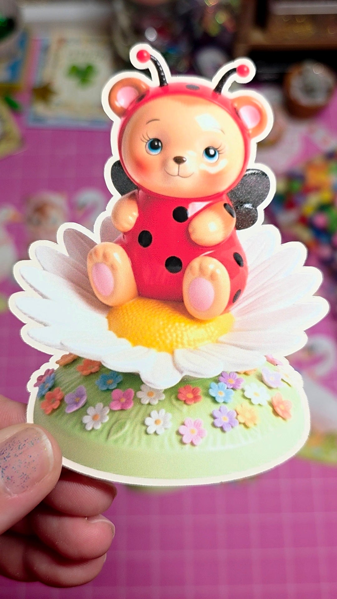 Cartoon ladybug character on a flower-shaped stand with colorful flowers around, held by a hand.