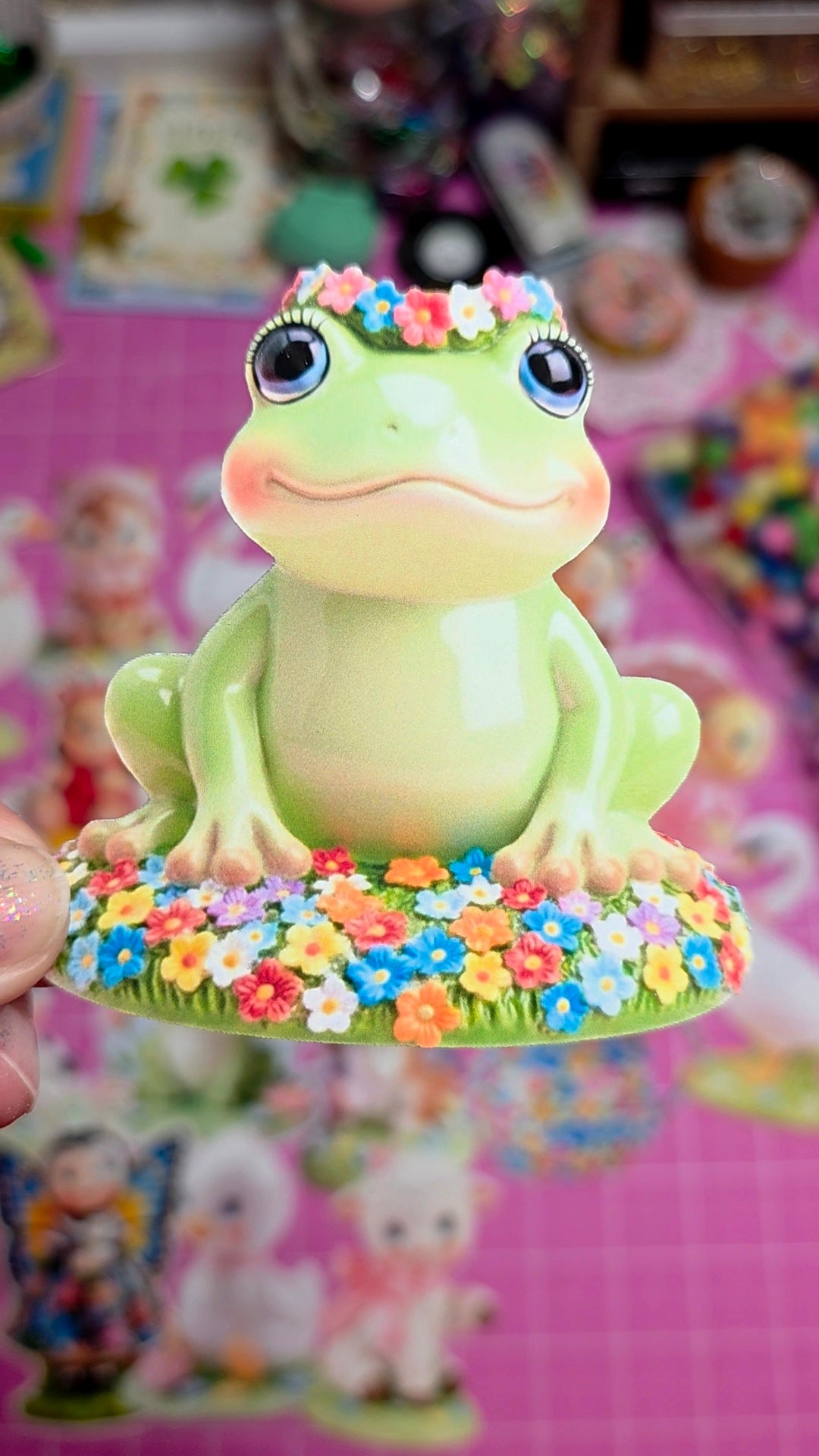 Decorative frog figurine with floral crown held against a colorful, blurred background