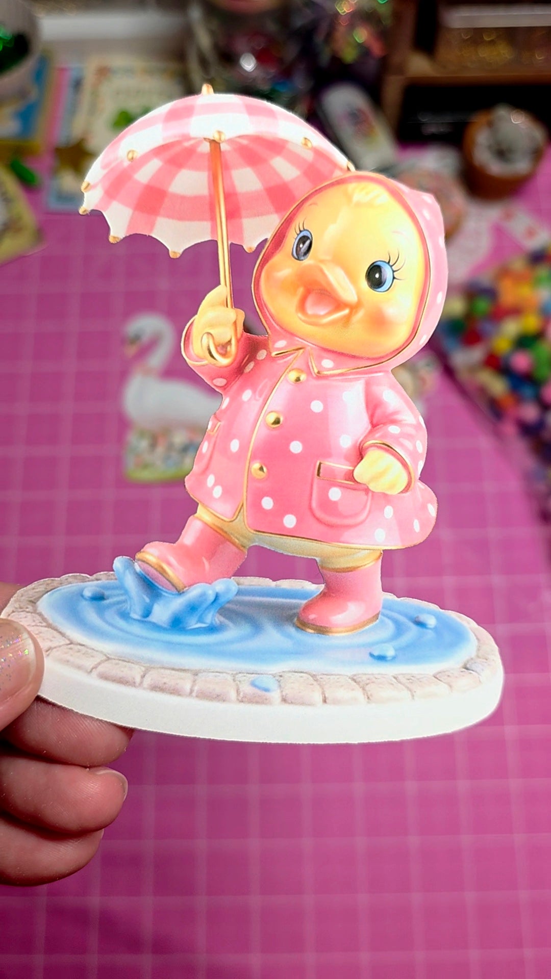Toy figure of a duck in a pink raincoat with an umbrella on a pink checkered background