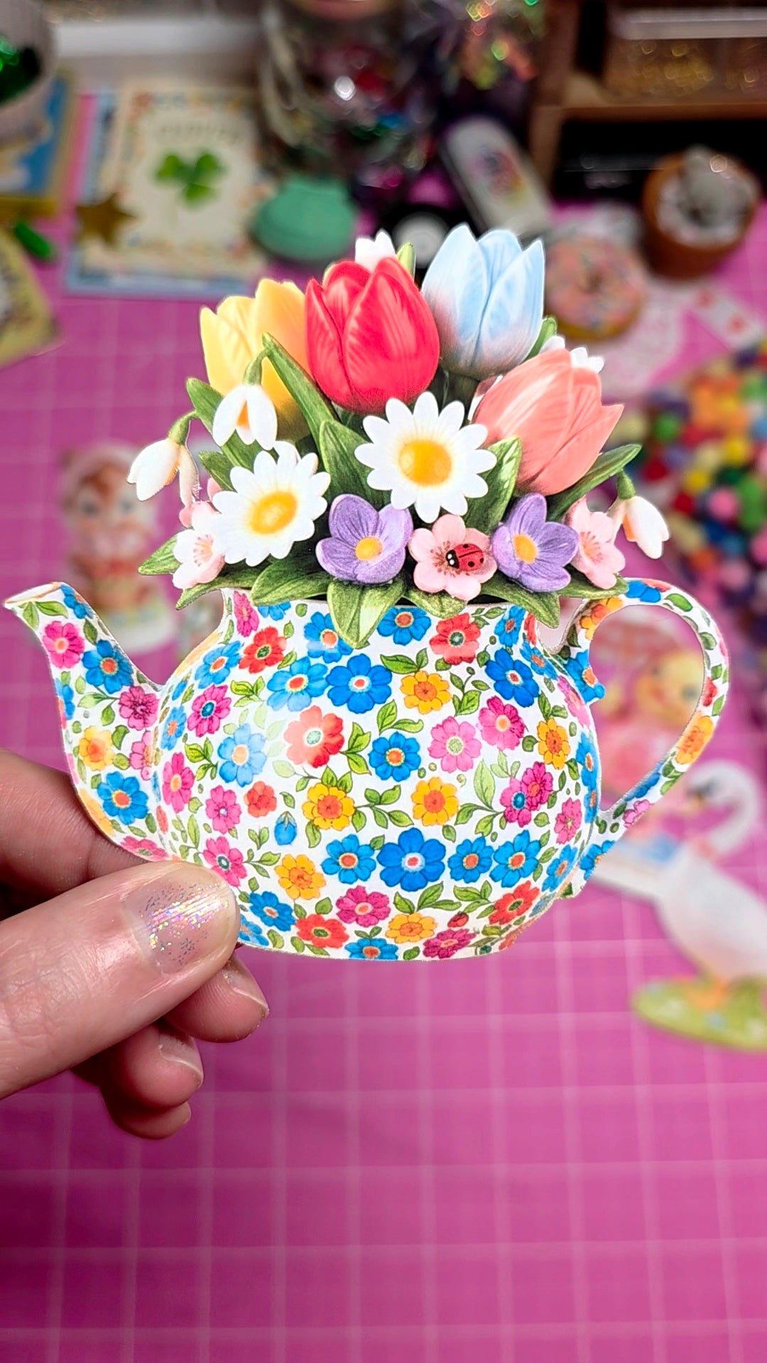 Colorful teapot-shaped flowerpot with flowers on a pink checkered background