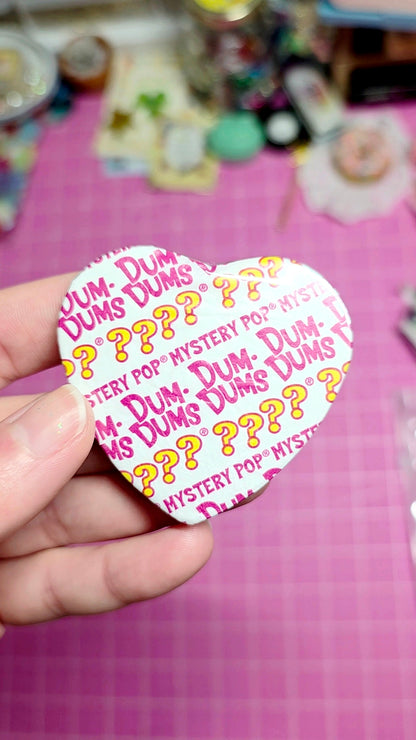 Heart-shaped Dum-Dums candy wrapper held by a hand on a pink surface.