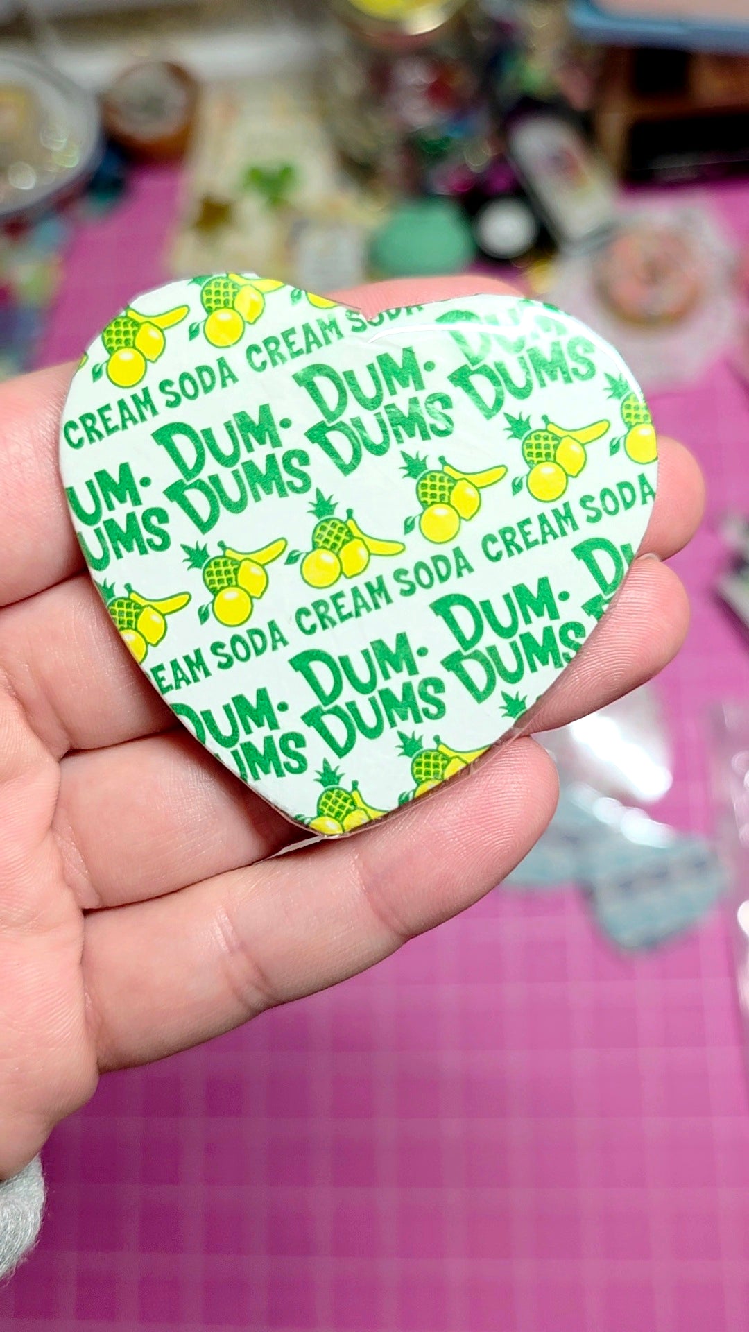 Dum-Dums lollipop container with green and yellow design held in a hand on a pink surface.