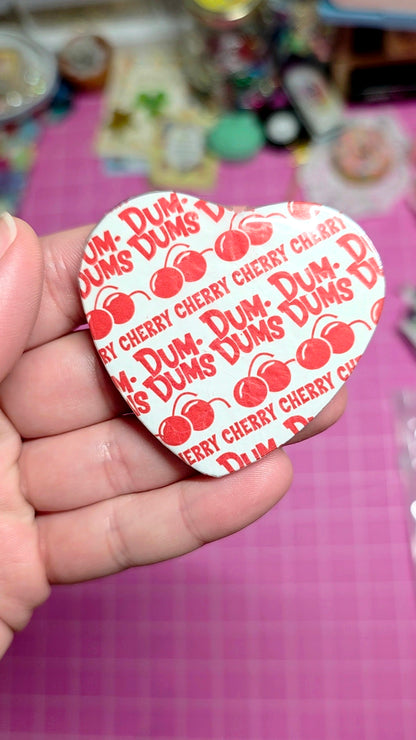 Heart-shaped Dum-Dum's candy wrapper held by a hand on a pink surface with blurred background