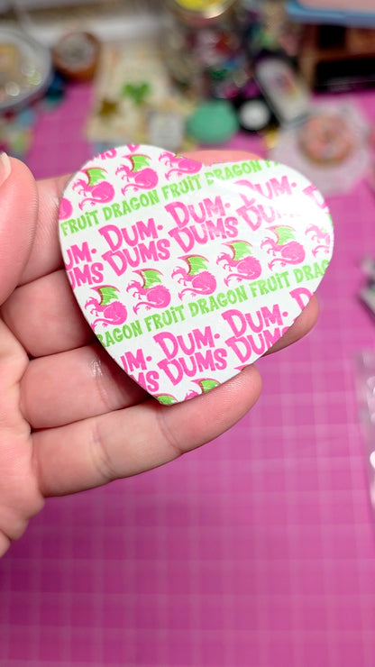Heart-shaped Dum-Dum's fruit dragon candy wrapper held by a hand on a pink surface.