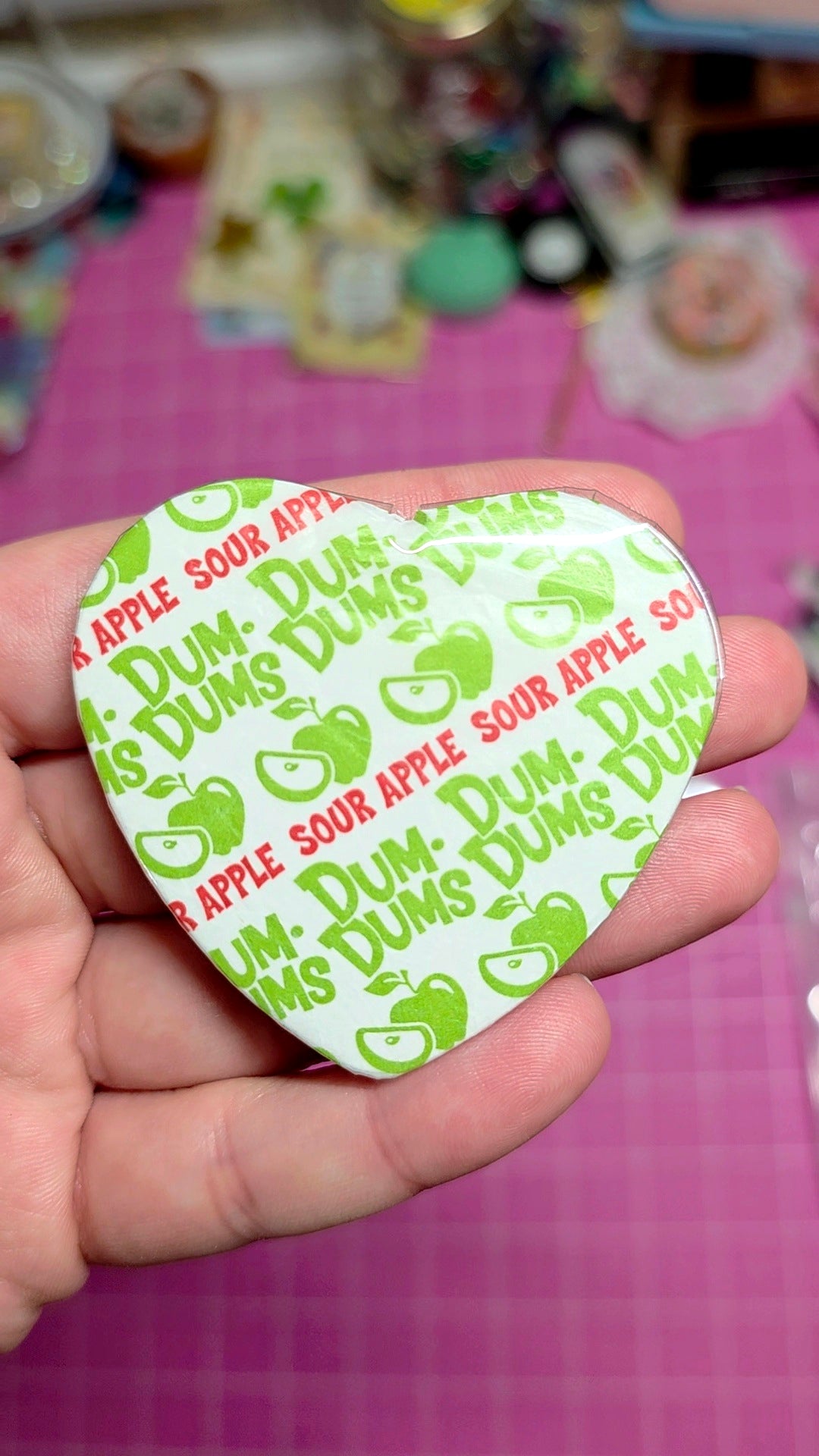Heart-shaped Dum Dums sour apple candy wrapper held in a hand with a colorful background.