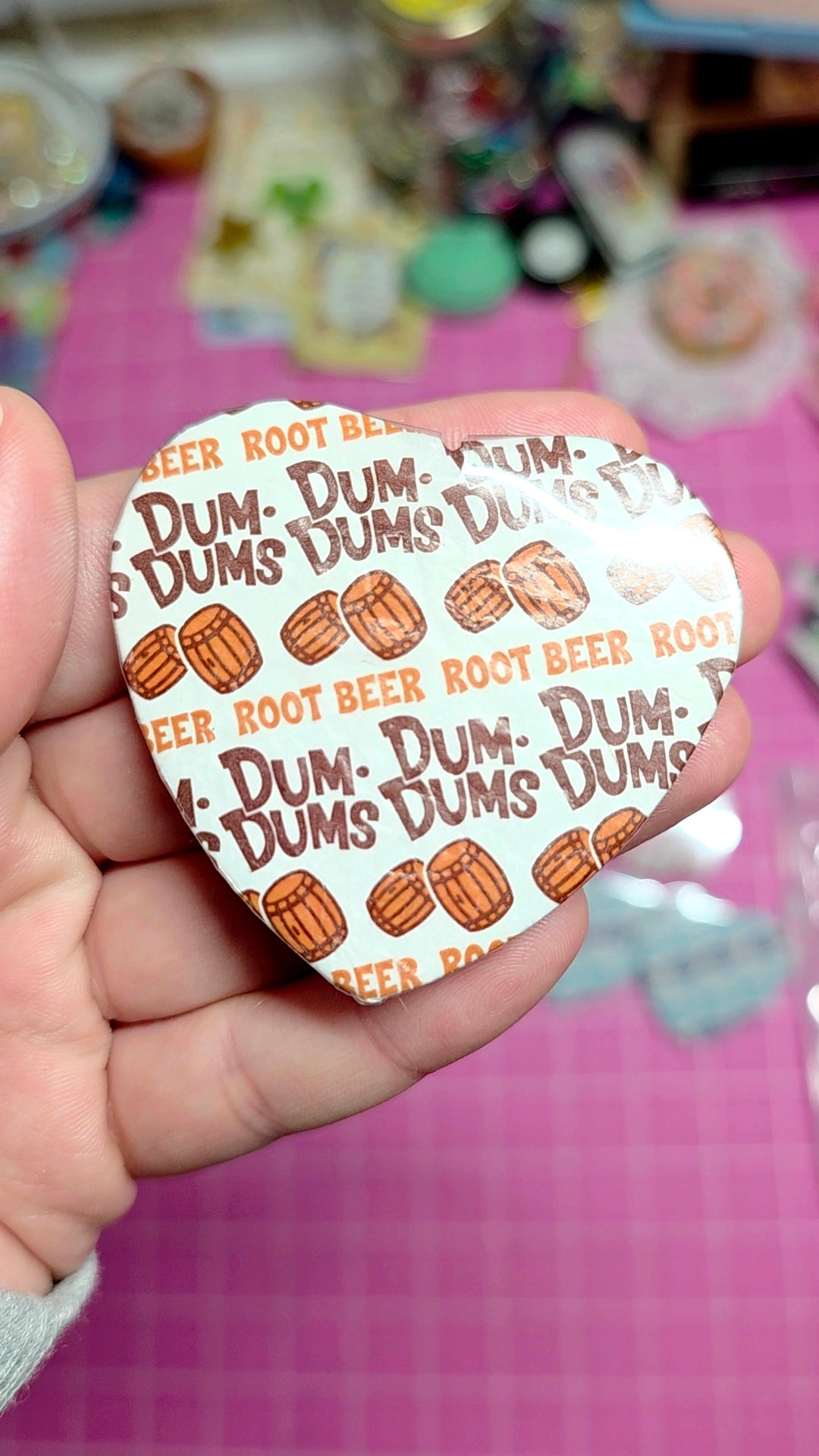 Heart-shaped sticker with Dum-Dum's Root Beer flavor text held in a hand against a pink background.